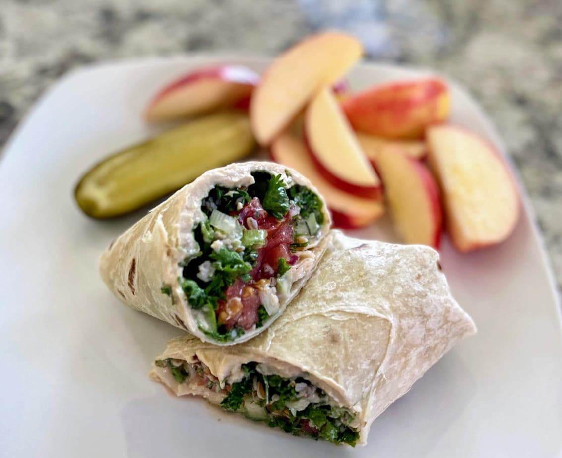 Hummus and Tabbouleh wrap served with sliced apples and pickles on a plate. 
