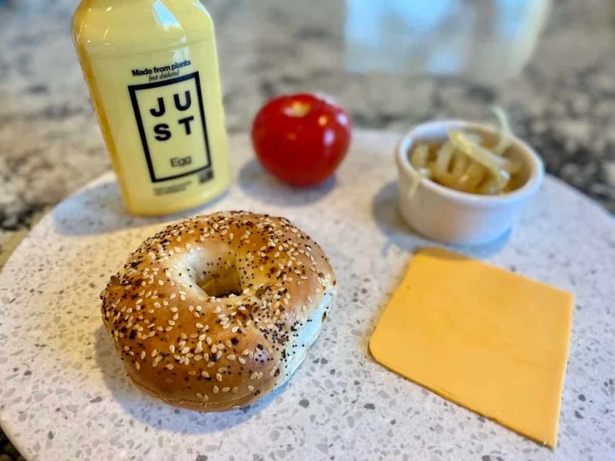 Breakfast sandwich ingredients on a cutting board: Just Egg, tomatoes, onions, cheese, bagel 