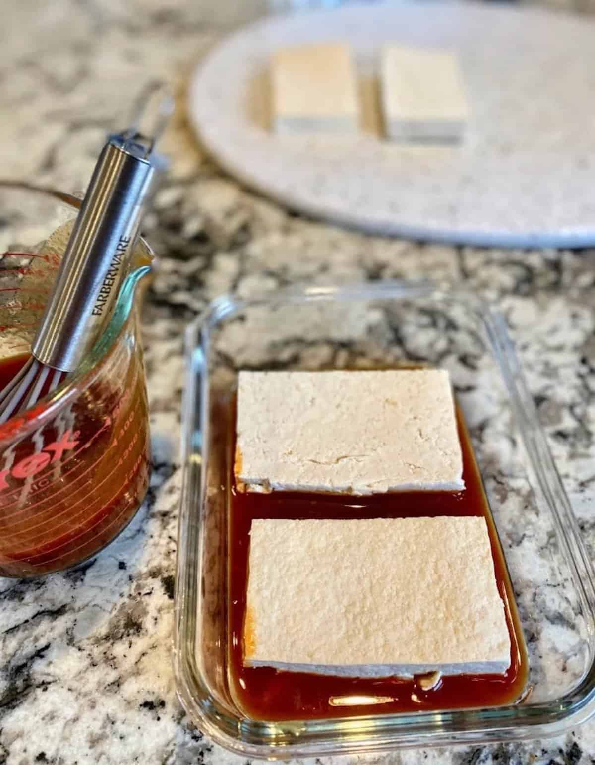 BBQ sauce inside a refrigerator safe container with labs of tofu to marinate on the counter.