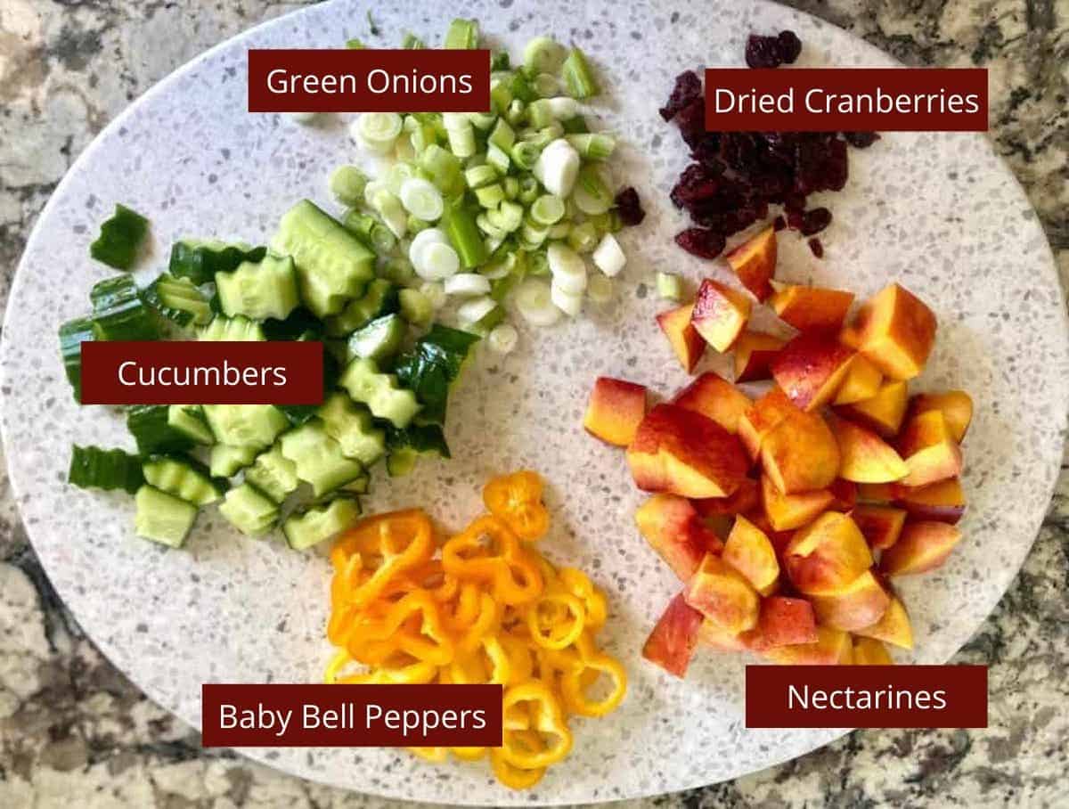 nectarine salad ingredients cut on a cutting board: green onions, dried cranberries, nectarines. baby bell peppers and cucumbers. 