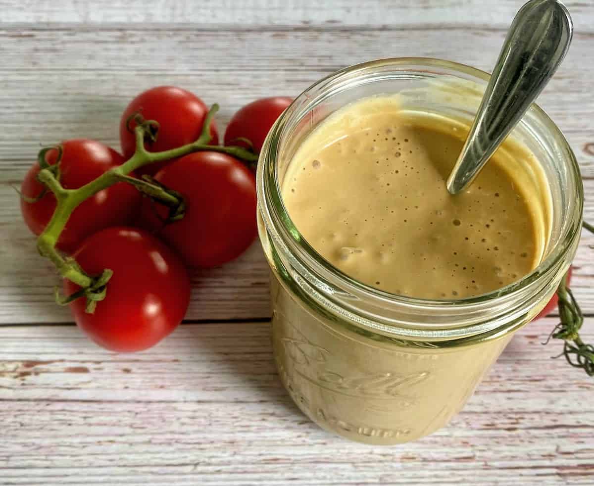 Creamy balsamic dressing is spooned into a bowl jar with cherry tomatoes on the table next to a vine.