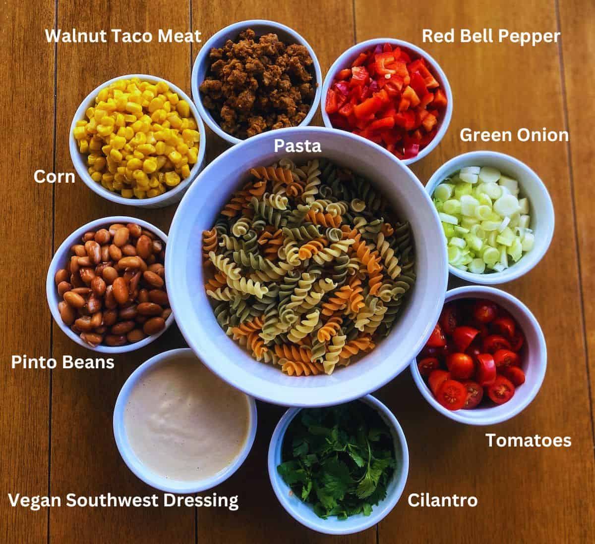 Taco pasta salad ingredients: Pasta, walnut taco meat, corn, pinto beans, green onions, tomatoes, cilantro, and vegan southwest dressing in bowls on the table