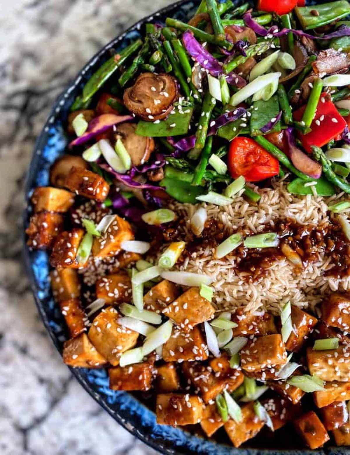 Kung Pau Tofu served with vegetables on top of brown rice drizzled with sauce. 