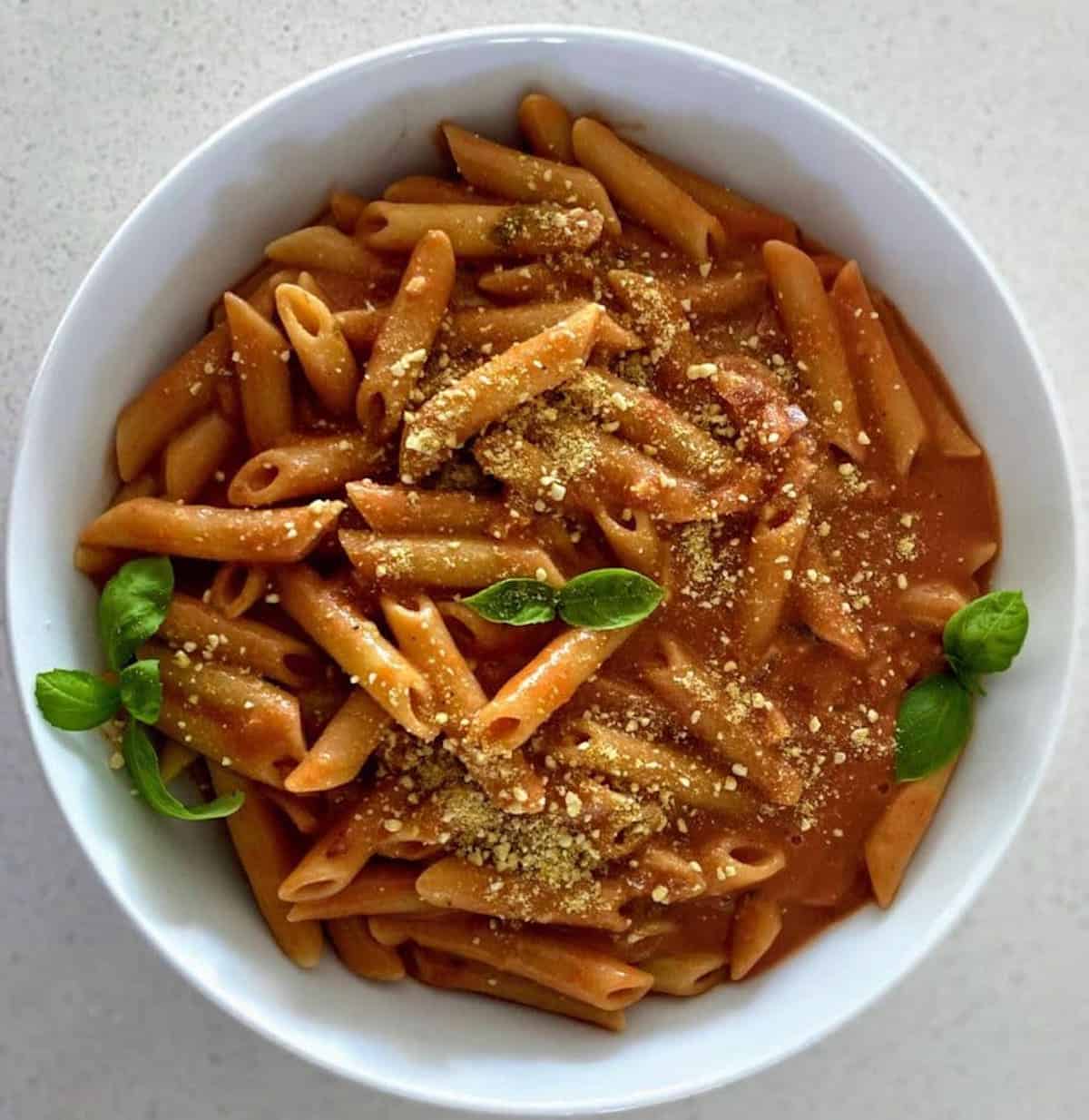 Penne Rose Pasta is in a bowl garnished with vegan parmesan cheese and fresh basil.