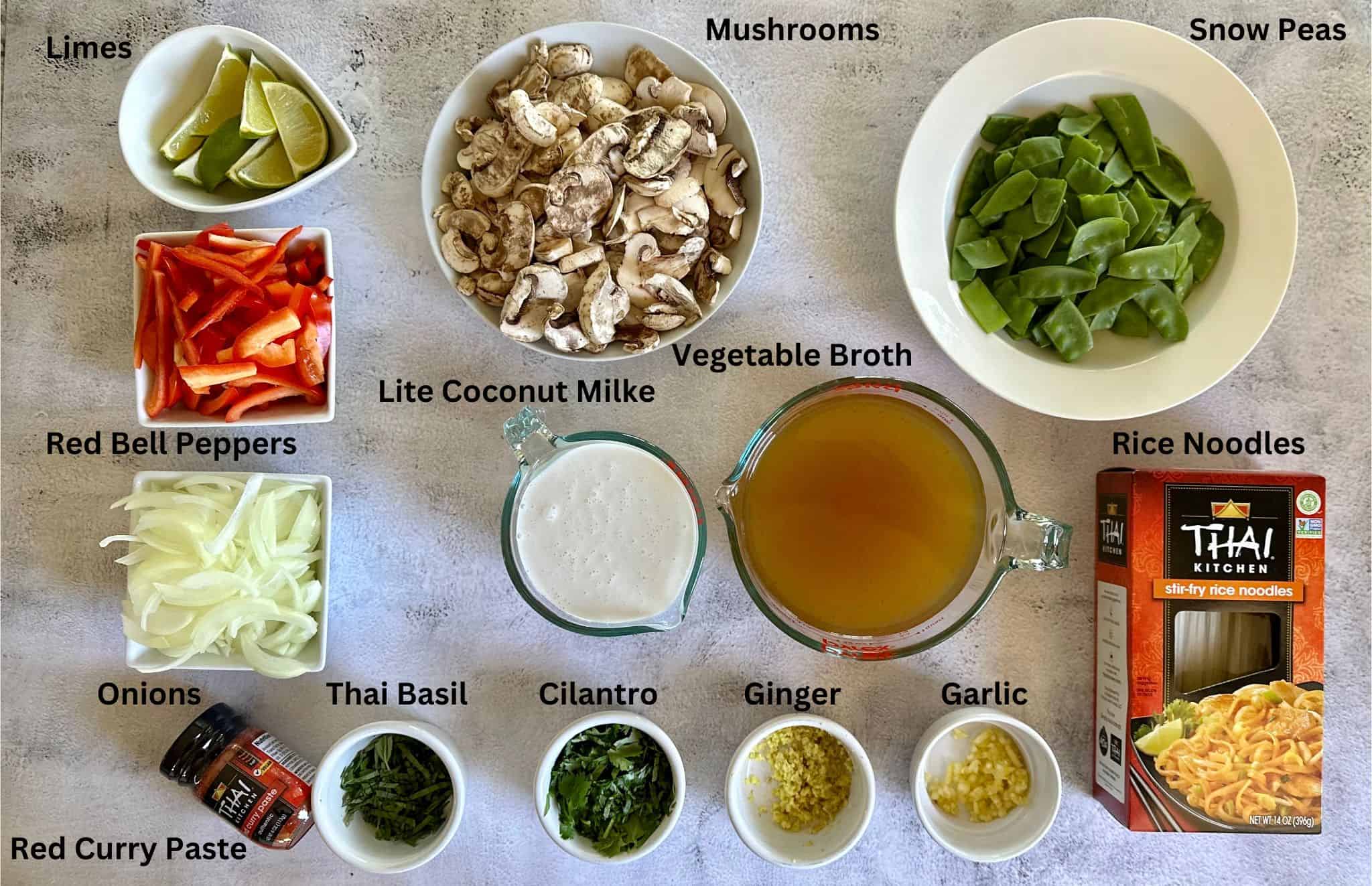 Recipe ingredients on the counter: rice noodles, Thai red curry paste, yellow onion, red bell peppers, mushrooms, snap peas, lite coconut milk, vegetable broth, ginger, garlic, cilantro, and Thai basil. 
