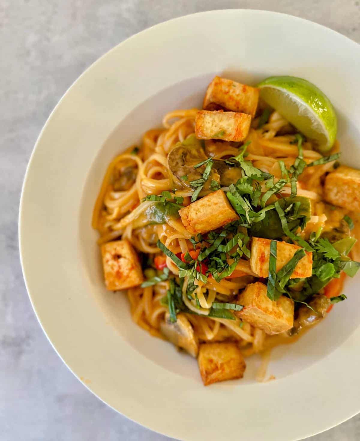 Red curry noodles in a bowl with tofu, Thai basil, and lime wedges. 