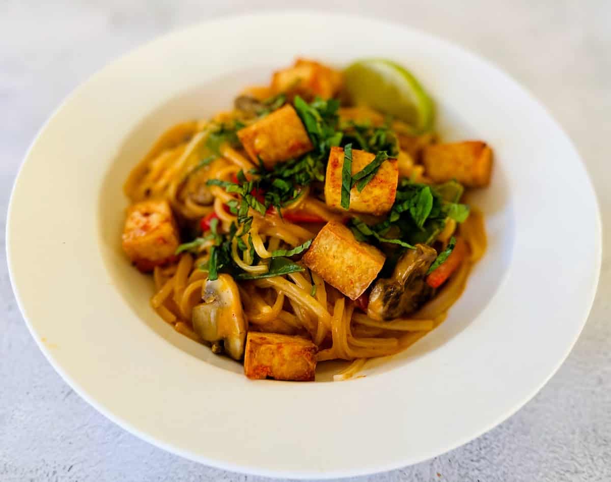 Thai Red Curry Noodles in a bowl garnished with curry tofu, cilantro, limes, and Thai basil.